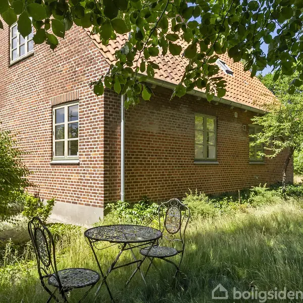 Rundt havebord og stole står på en græsplæne ved et rødt murstenshus med vinduer. Træer og buske omkranser huset i en frodig have.