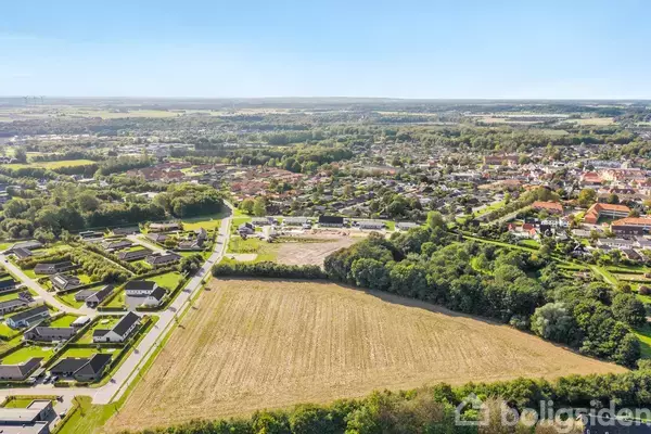 En landbrugsmark ligger stille omgivet af huse og træer i en by. Landskabet strækker sig ud mod horisonten under en klar blå himmel.
