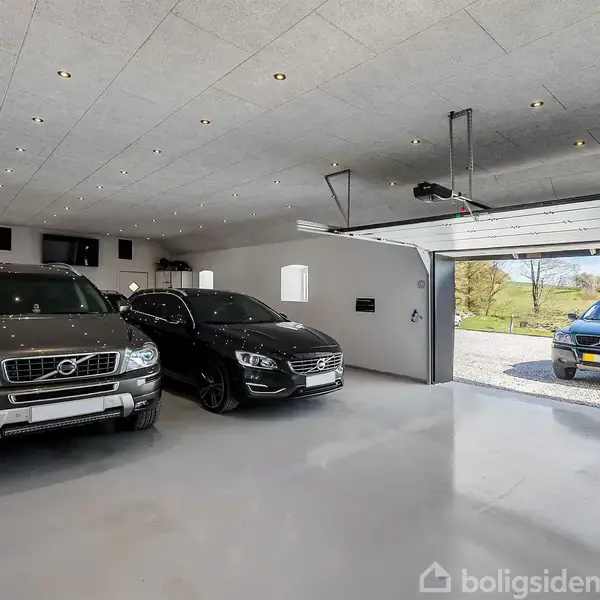 To biler holder parkeret i en rummelig garage med åbne døre, mens en blå bil står udenfor på en grusvej. Garagen er lyst op og moderne indrettet.