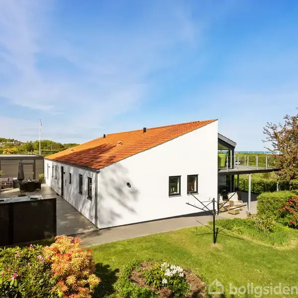 En hvid, moderne villa med rødt tegltag står i en velplejet have, omgivet af blomstrende buske og grønne planter under en klar blå himmel.