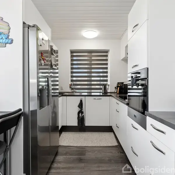 Køkken med hvide skabe, sort bordplade og mørkt gulv. Et køleskab står ved siden af et rundt bord med en stol. Et lille vindue lukker lys ind. En vægdekoration siger "Home Sweet Home".