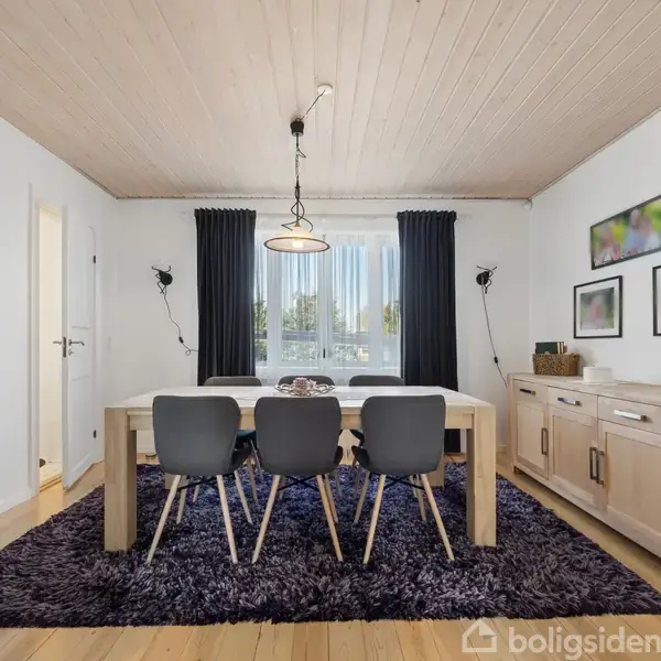 Spisebord med fire stole står midt i et lyst rum med træloft. En lampe hænger over bordet. Et skab står til højre, og der er hvide vægge med billeder.