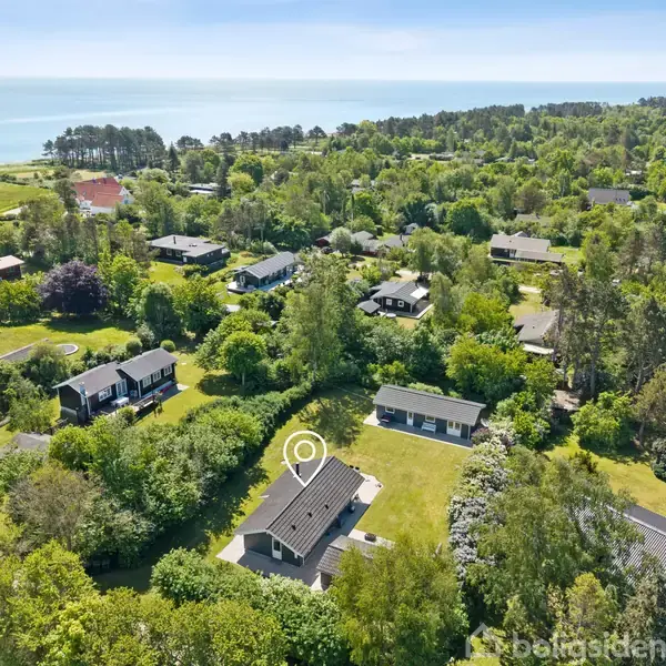 Et hus er markeret med en rund cirkel i en frodig, skovfyldt landsby nær kysten. Flere huse og træer omgiver området med havet i baggrunden.