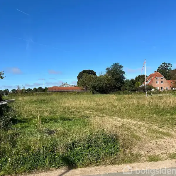 En flagstang står i en grøn eng under en klar blå himmel. I baggrunden ses et rødt murstenshus med træer omkring.
