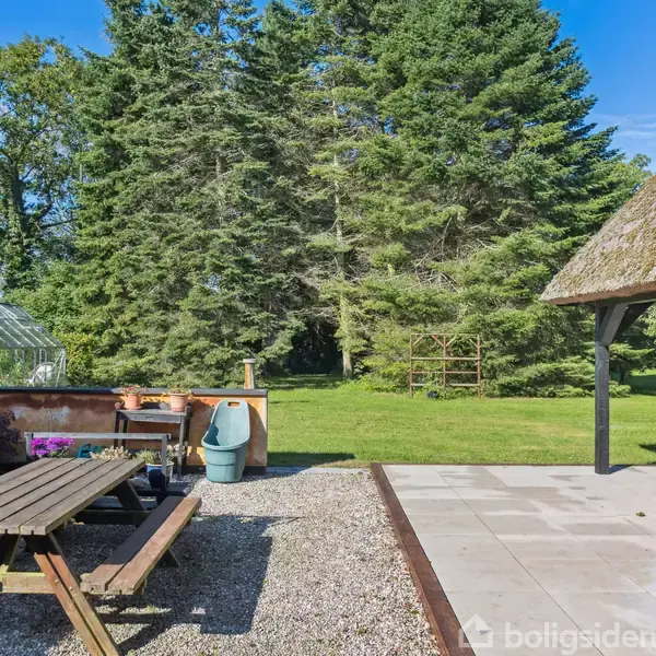 Et stråtækt hus med åbent terrasseområde, hvor der står borde og bænke. Omgivet af en grøn græsplæne og træer i baggrunden under en blå himmel.