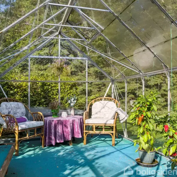 Træbænk står stille ved siden af bord på grusområde; omgivet af orange bygninger, græsplæne og skov med tætbevoksede træer i baggrunden. En glaspavillon ses til venstre.