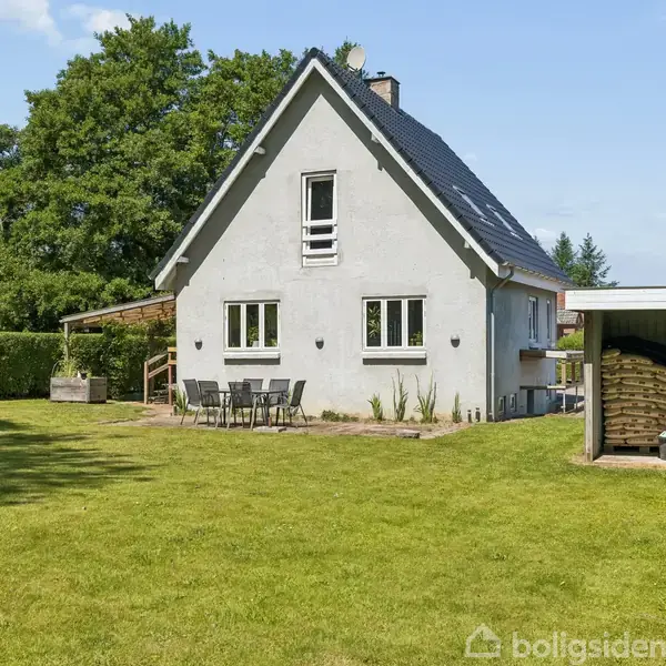 Hvidt hus med stejlt tag i en grøn have. Foran huset står et bord og fire stole. En overdækket brændestabel ses til højre, omgivet af træer.