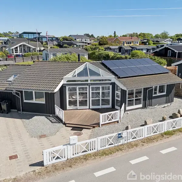 Et enfamiliehus med mørke facader og solpaneler på taget står bag et hvidt stakit. Huset har store vinduespartier og ligger i et villakvarter med blå himmel.