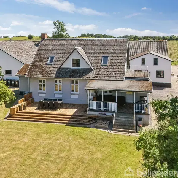 Et hus med en stor træterrasse står midt i en grøn have, omgivet af landbrugsbygninger. Terrassen har udendørs møbler, og der er en overdækket veranda.
