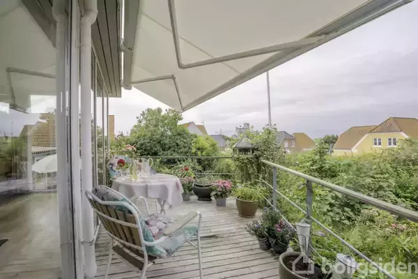 Terrasse med bord og stole er indrettet til hyggelig udendørsspisning. Omgivet af planter og blomster i krukker. Udsigt til nabolagshuse og grønne områder under en stor markise.