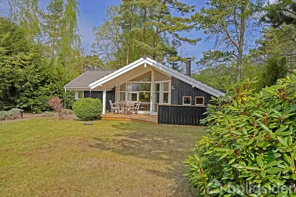 Sort træhus med store vinduer og overdækket terrasse, omgivet af græsplæne og høje træer. Terrasse har et bord med stole, tilbyder skyggefuldt opholdsområde i skovagtig have.