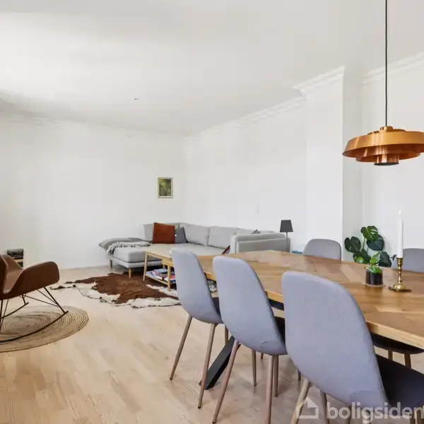 Spisebord med grå stole står i lys stue med trægulv. I baggrunden er en grå sofa og tæppe på gulvet. En kobberlampe hænger over bordet.