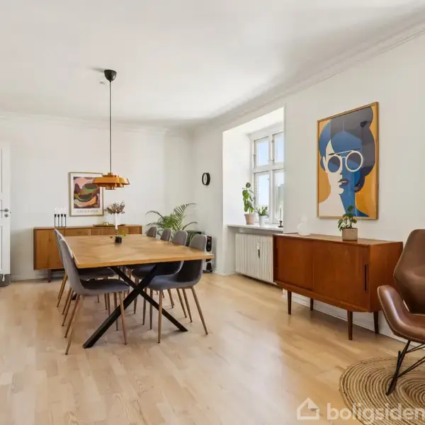 Spisebord med stole omkring; står centralt i en lys stue; omgivet af moderne kunst, planter og træmøbler, herunder en lav reol under vinduet og en lænestol i hjørnet.