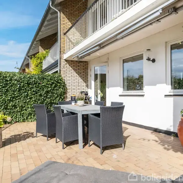 Et udendørs spisebord med seks sorte stole står på en terrasse med gule fliser. Vegetation og murstensvægge omkranser området. Skyfri blå himmel danner en lys og åben atmosfære.