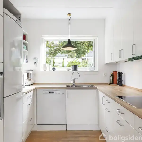 Køkken med hvide skabe og indbyggede apparater; en ovn og opvaskemaskine. Et stort vindue til venstre slipper lys ind, mens en loftslampe hænger over vasken.