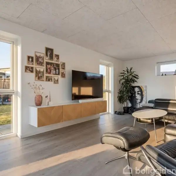 Et moderne stueområde med en stor sort lædersofa, et vægmonteret TV, og et sofabord. Væggene har billedrammer og en plante står ved siden af sofaen. Udsigt til have.