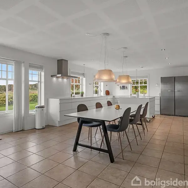 Et moderne køkken med et langt spisebord og flere stole. Store vinduer giver udsigt til en grøn have. Lyse lamper hænger over bordet, skaber en lys og åben atmosfære.