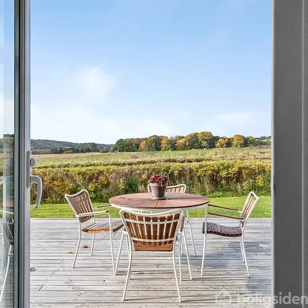 Rundt havebord med fire stole står på en træterrasse. En blomsterpotte pryder bordet. Udsigt over en frodig mark med skov i baggrunden ses gennem en døråbning.