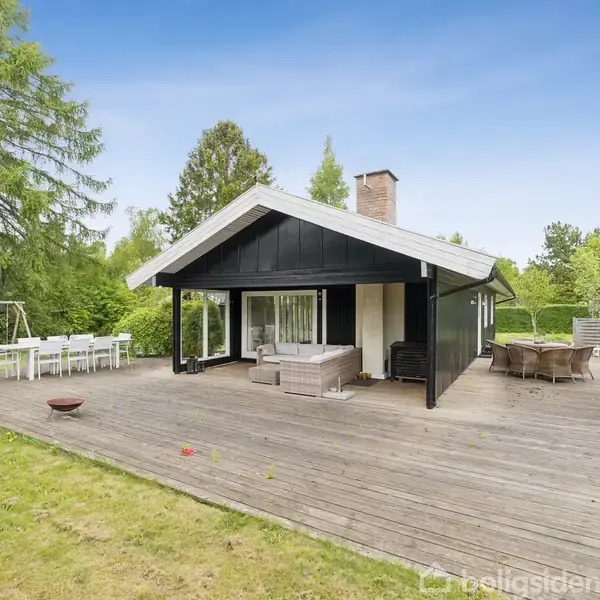 Et sortmalet sommerhus med terrasse i træ omgivet af frodig have. Terrasse med siddemøbler og spiseområde. Store træer og buske i baggrunden under en klar blå himmel.