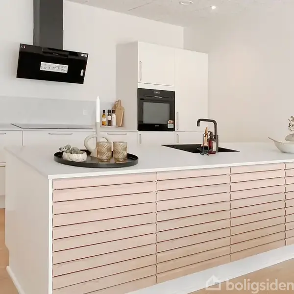 Et moderne køkken viser en køkkenø med hvide skuffer og en indbygget vask. En emhætte og en indbygget ovn ses i baggrunden. Rummet er lyst og minimalistisk.