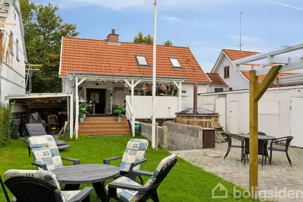 Et rødt taghus har en overdækket terrasse med planter, omgivet af stoler med hynder i en græsplæne og en flisebelagt område med bord og stole.