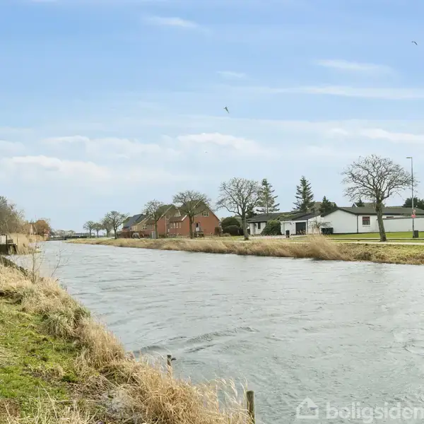 Flod løber roligt gennem et landskab med rand af græs og buske. I baggrunden ses få huse og træer under en klar blå himmel.