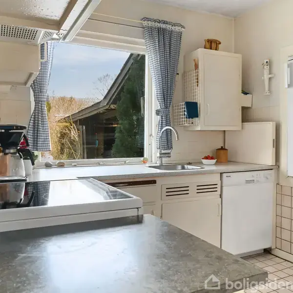 Et køkken med komfur og kaffemaskine på en bordplade, ved siden af et vindue med udsigt til en have. Hvide skabe og fliser omkranser området. En stol ses til højre.