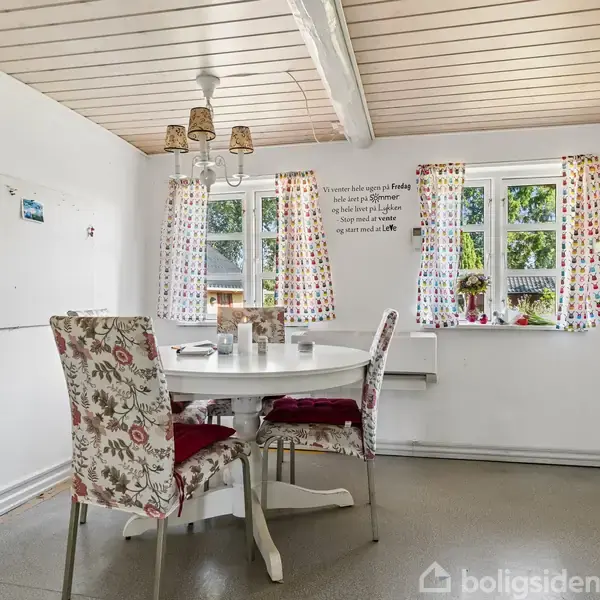 Rundt bord med fire blomstrede stole står i et lyst køkken. Vinduer med farverige gardiner slipper lys ind. Skilt på væggen siger: "What matters most is Family".