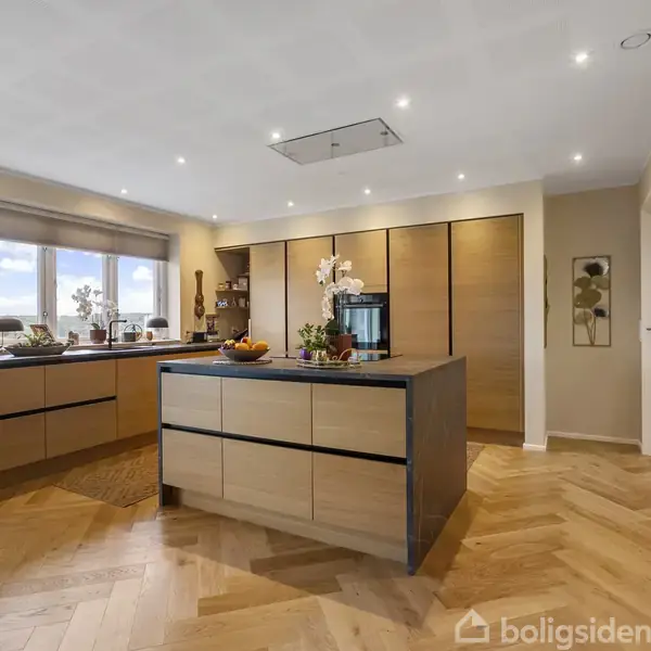 Et moderne køkken med ø, hvorpå der står frugtskål og planter. På væggen ses teksten "The kitchen is the heart of this house." Rummet har trægulv og store vinduer.