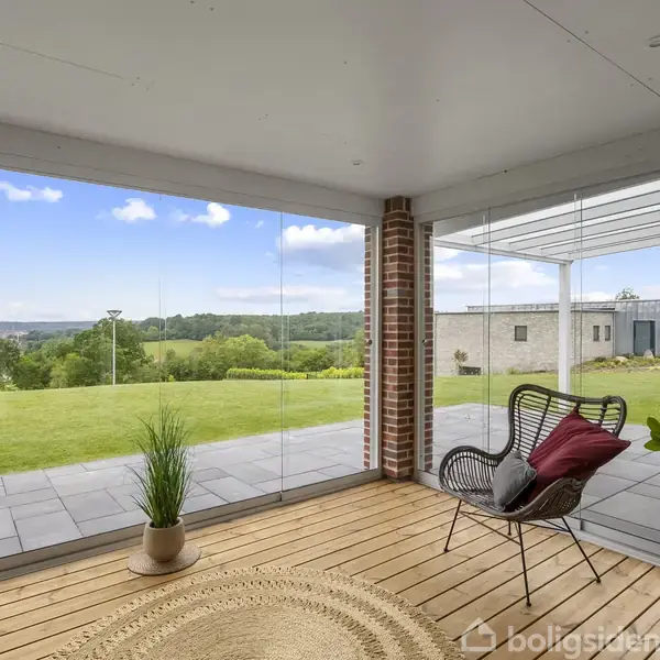 Overdækket terrasse med en moderne stol og pude, planter og et rundt tæppe. Skydedøre åbner til en velplejet græsplæne med landskabsudsigt. Klare glasvægge giver uhindret udsigt over området.