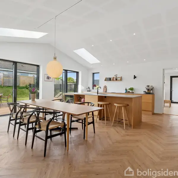 Spisebord med stole står centralt på trægulv i en moderne, lys stue. Køkkenområde med barstole i baggrunden. Store vinduer og glasdøre fører til en have.