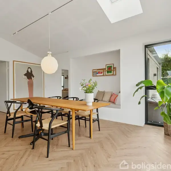 Et spisebord i træ er omgivet af sorte stole. Rundt om bordet hænger en hvid pendellampe. Rummet har lyst parketgulv, store vinduer, en sofa og planter.