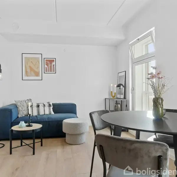 En stue med en blå sofa står ved en hvid væg med to kunstværker. Et rundt bord med stole og en vase med blomster står i forgrunden.