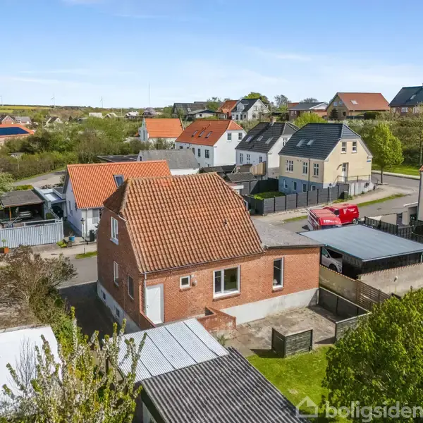 En rødstensvilla med rødt tegltag står stille i et boligkvarter med flere lignende huse. Flere af husene har hvide vinduesrammer, og en rød bil er parkeret ved vejen.
