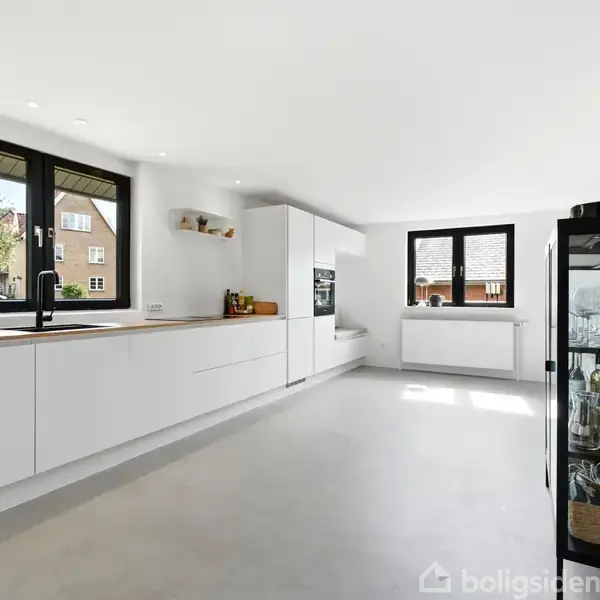 Et moderne køkken har hvide skabe og en sort glasvitrineskab. Det er indrettet i et lyst rum med store vinduer, der giver udsigt til det omkringliggende boligområde.