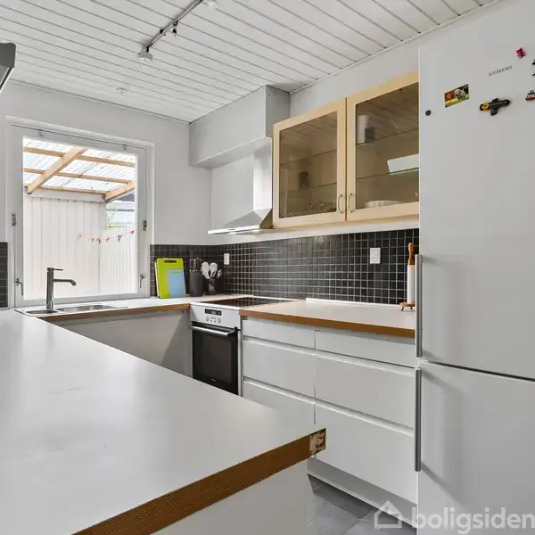 Et lyst køkken har hvide skabe og en hvid bordplade. En opvaskemaskine er under bordet ved vinduet. En køleskab med magneter står ved siden af.