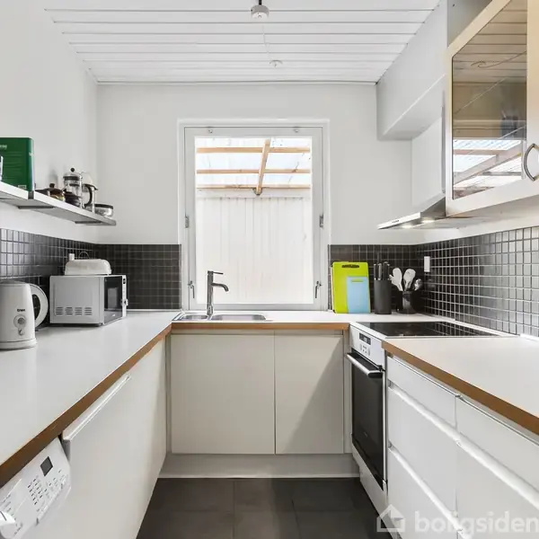 Et moderne køkken med hvide bordplader, hvor en elkedel og mikroovn står på bordet. Sorte fliser pryder væggene, og et vindue giver naturligt lys. Hylder indeholder kopper og glas. Ingen tekst er til stede.