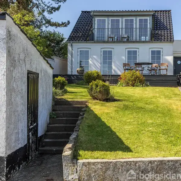 Hvidt hus med sort tag står på en hævet terrasse. Et bord og stole er udenfor. En græsplæne omkranser stentrapperne, der fører op til huset, med buske ved siden.