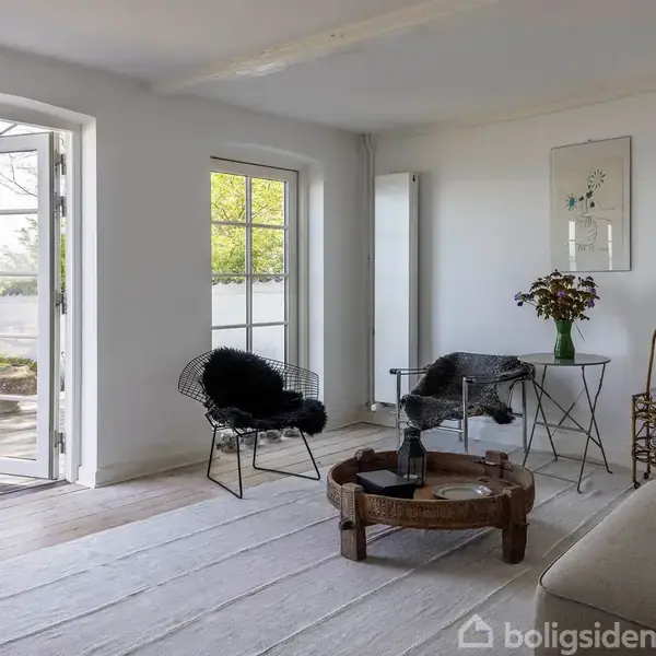 Stue med sofa til højre, to stole og et rundt træbord. Dør og vindue åbner mod en terrasse med udsigt til have. Blomster står på et bord ved væggen.