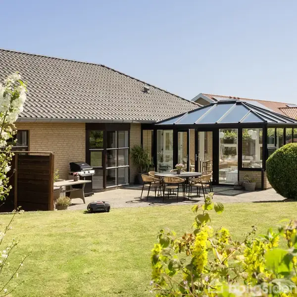 Rødstenshus med terrasse og glaskarnap omgivet af velholdt have. Bord og stole står på terrassen, mens grillen står til venstre. Husene i baggrunden skaber et nabolagsmiljø.