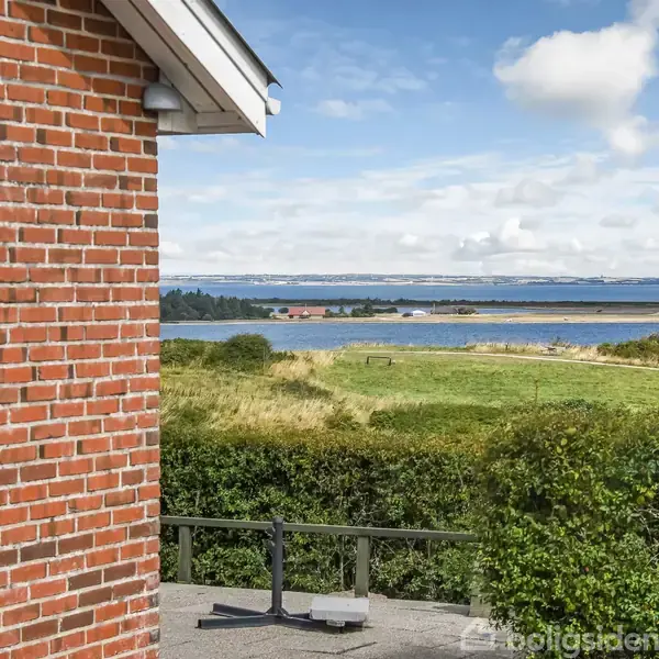 En del af et murstenshus står stille ved siden af en terrasse med udsigt over en frodig eng og en fjord i det fjerne under en blå, skyet himmel.