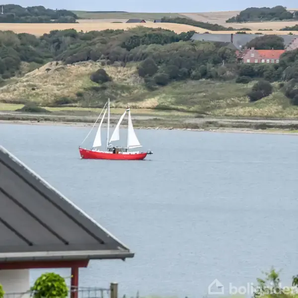 En rød sejlbåd bevæger sig på en sø omgivet af bakker og huse. I forgrunden ses et tag og buske.