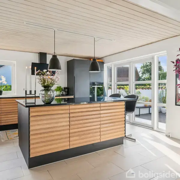 Køkken med træskabsfronter og sorte hængelamper, stående i dagslys fyldt rum. Planter og blomster dekorerer rummet, mens et stort vindue afslører en terrasse udenfor.