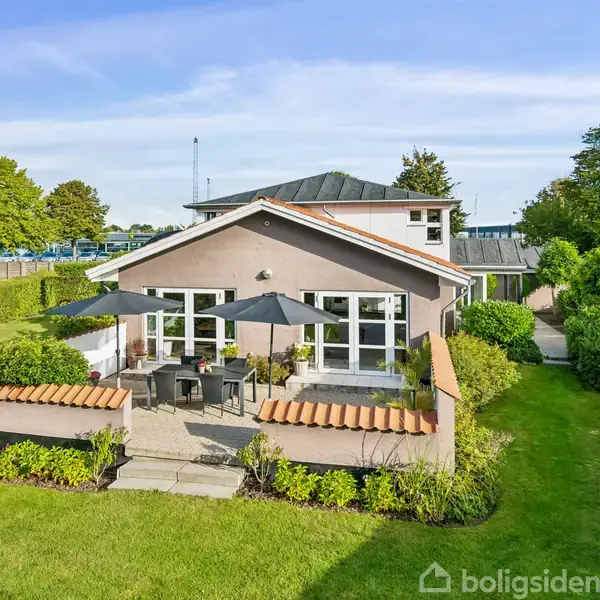 Et hus med moderne arkitektur står fredeligt i en velplejet have. To parasoller skygger over havemøbler på terrassen, omgivet af grønne buske og træer i et solrigt område.