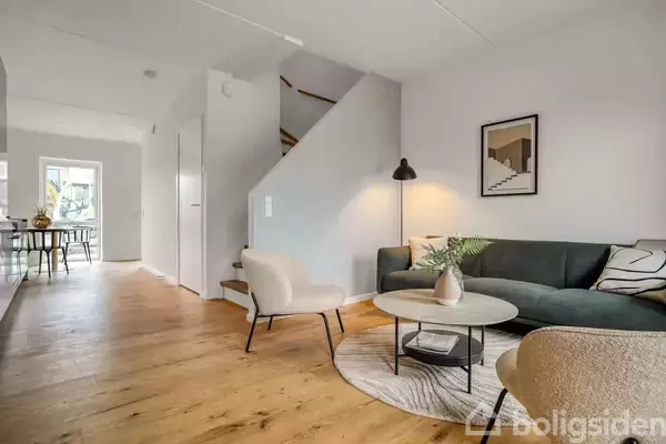 Stue med mørkegrøn sofa, sofabord og gulvlampe, placeret på lyst trægulv; i baggrunden ses en spisekrog ved vinduet og en trappe op ad væggen.