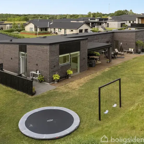 Moderne, sort murstenshus står i en velholdt have med en trampolin og gyngestativ. Flere husdyrkrukker pryder terrassen, hvor der er opsat havemøbler og grilludstyr.