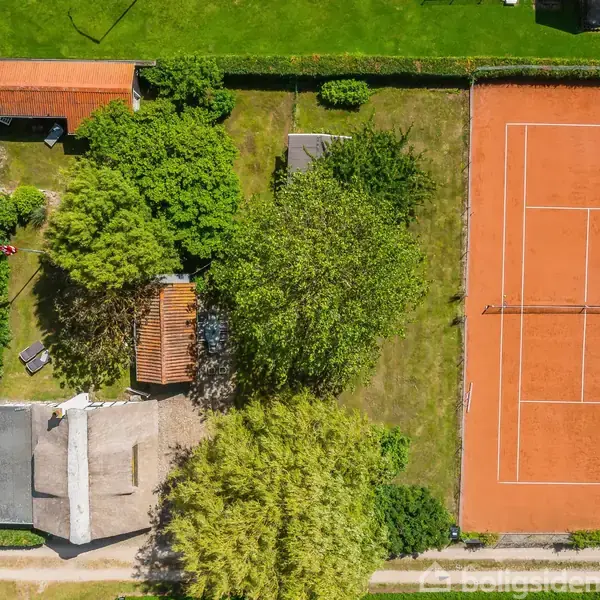 Tennisbane med rød jordoverflade omgivet af hække. Ved siden af ses en have med flere træer og en bygning med rødt tag. Grøn græsplæne omkranser området.