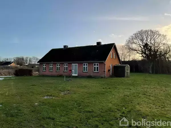 Et rødt murstenshus med sort tag står på en stor, grøn græsplæne. Baggrunden har træer og en klar himmel.