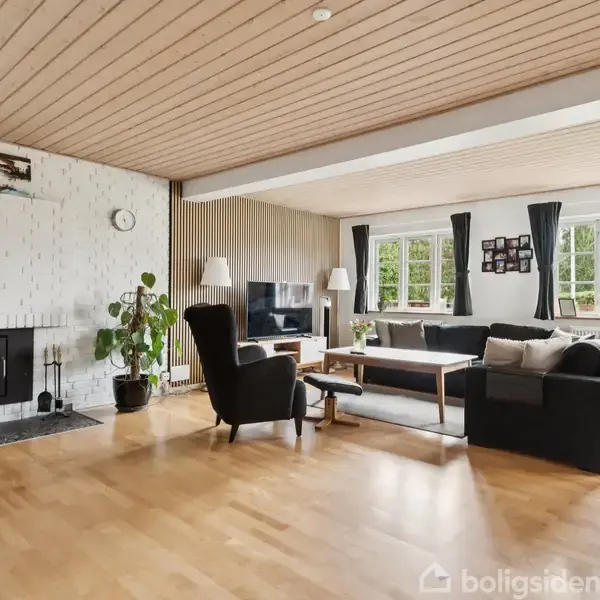 Stue med sofa og lænestole omkring et sofabord, fjernsyn ved væggen. Brændeovn til venstre med grøn plante ved siden, træloft og parketgulv. Store vinduer med gardiner.