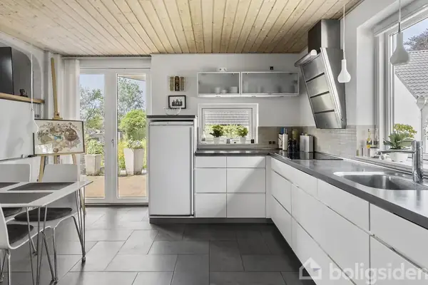 Et moderne køkken med hvide skabe, stål-emhætte og grå fliser. Bordet har sorte underlag, og en dør fører ud til en terrasse med planter. Vinduet slipper lys ind.